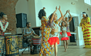 A group of people wearing colorful clothing dance together, some with their arms raised above their heads